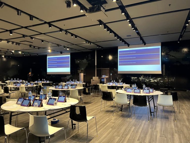 A modern conference room set up with round tables, individual digital tablets, and large projection screens, demonstrating the logistical complexity of why biotech events require planner services.