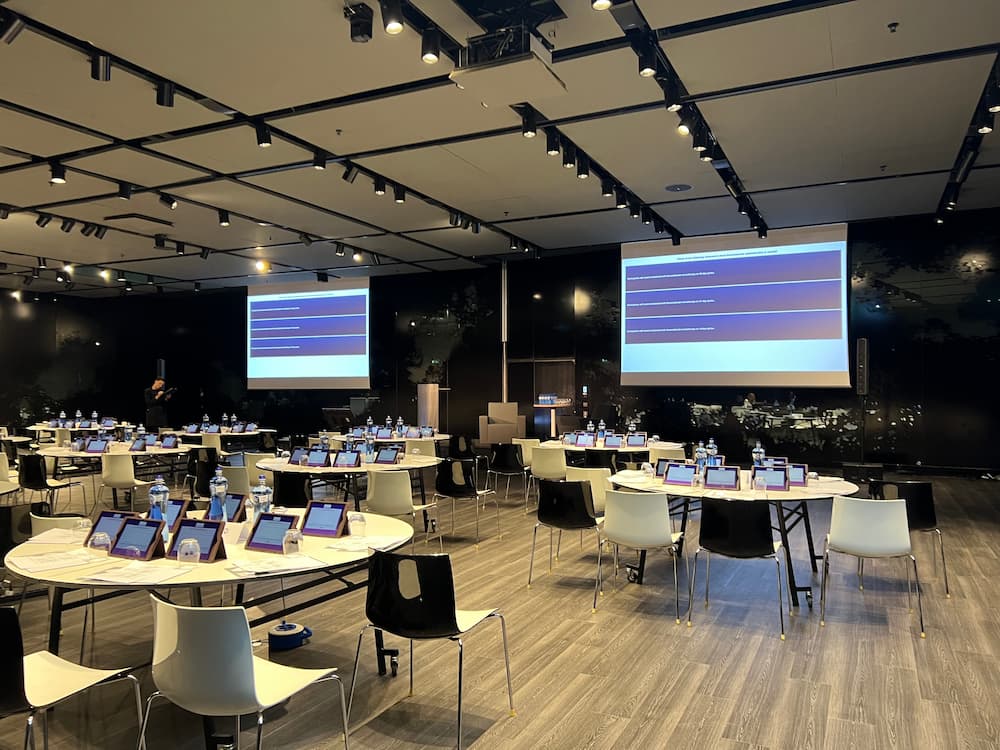A modern conference room set up with round tables, individual digital tablets, and large projection screens, demonstrating the logistical complexity of why biotech events require planner services.
